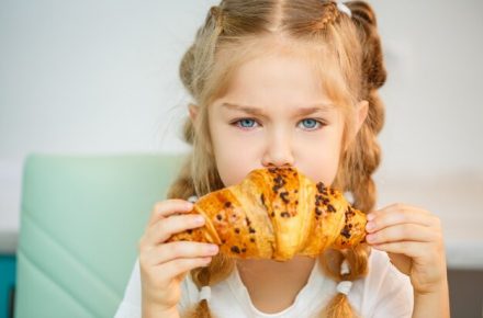 portrait-little-blonde-woman-sitting-kitchen-biting-fresh-croissant_173815-11980